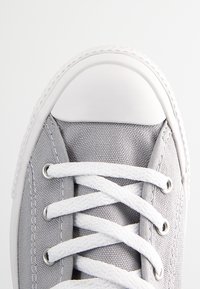 Szare płócienne trampki z białym gumowym noskiem i oczkami, z białymi sznurowadłami przewleczonymi przez srebrne oczka i teksturowaną powierzchnią.