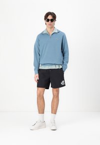 Hanorac bleu deschis cu guler cu fermoar, căptușeală cu dungi, pantaloni scurți negri, adidași albi și ochelari de soare. Prezintă un logo pe piept.
