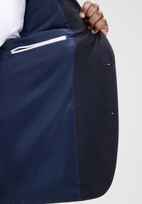 Innenansicht eines marineblauen Blazers mit glattem Futter, das eine weiße Tasche, schwarzen Stoff mit Steppnähten und zwei kleine seitliche Öffnungen zeigt.