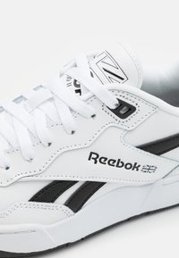 Adidași din piele albă cu accente negre, având un design cusut, șireturi albe groase și un logo Reebok pe lateral.