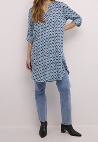Jasnoniebieska tunika z czarnym falistym wzorem, dekoltem w serek i rękawami o długości trzy czwarte, zestawiona z jasnoniebieskimi dżinsami i czarnymi botkami na obcasie.