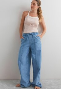 Jeune femme avec de longs cheveux en queue de cheval portant un débardeur rayé blanc et orange et un jean large bleu, pieds nus, adossée à un mur.