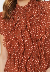 Ruftad blus i bränd orange med vita prickar; har hög hals och knäppning, tillverkad av lätt, texturerad textil.