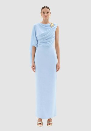 Donna che indossa un abito da sera azzurro chiaro monospalla con tessuto drappeggiato e una spilla dorata, abbinato a sandali dorati con tacco alto.