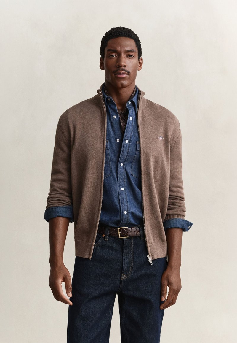 Homme portant un cardigan marron zippé sur une chemise en jean bleue boutonnée et un jean foncé, debout devant un fond clair uni.