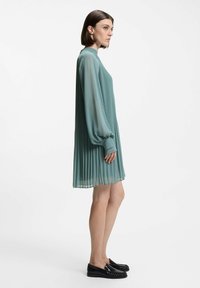 Pleated, teal dress with a high neckline and long sleeves. Fabric is lightweight and flowing. Paired with black loafers.