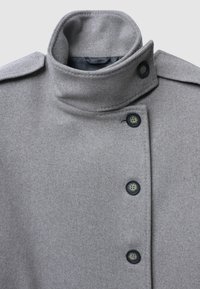Manteau en laine gris avec un col montant, présentant un design croisé à double boutonnage et de grands boutons noirs, une texture lisse et des accents minimalistes.