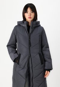 Manteau matelassé à capuche, long et gris foncé, avec un biais en chevron, fermeture éclair et poches latérales. Il a des poignets élastiques et un tissu texturé.