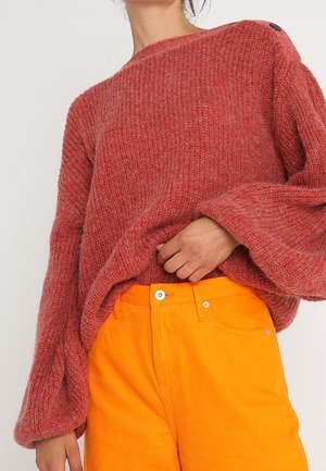 Loszittende, getextureerde rode trui met lange, wijde mouwen, gedragen in een feloranje broek met hoge taille en knoopsluiting.