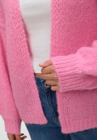Κλείσιμο εικόνας ενός ατόμου που φοράει μια ροζ, χνουδωτή πλεκτή ζακέτα πάνω από ένα λευκό τοπ και μπλε τζιν, κρατώντας το τελείωμα της ζακέτας με το ένα χέρι.