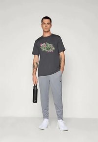 Camiseta gráfica de Nike en gris oscuro con logo colorido, combinada con pantalones deportivos gris claros, y sosteniendo una botella de agua negra. Zapatillas blancas.