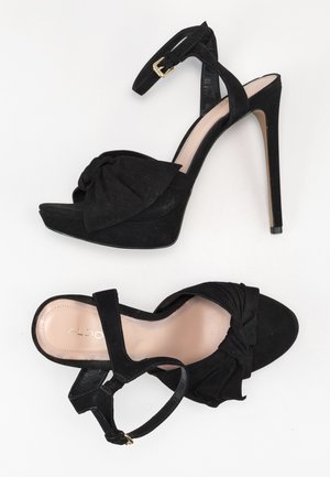 Czarne zamszowe sandały na wysokim obcasie z grubą platformą, paskiem wokół kostki i węzłowym detalem na przodzie. Złota klamra na pasku.