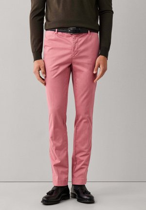 Man wearing slim-fit pink trousers, black belt, black tassel loafers, and a dark long-sleeve top standing against a plain background.
