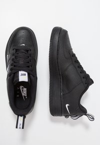 Černé tenisky Nike Air s texturovaným koženým povrchem, šněrovacím designem, perforovanou špičkou a bílými logovými akcenty na stranách a jazyku.