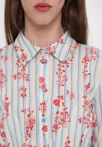 Cvetna košulja na gumb z bledo podlago, rdečimi in modrimi navpičnimi črtami, z majhnimi rdečimi cvetovi in svetloblue gumbi.