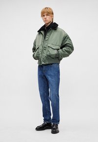 Chaqueta bomber verde con cuello de piel negra, bolsillos con cremallera, combinada con vaqueros azules y zapatos negros, posando frente a un fondo liso.