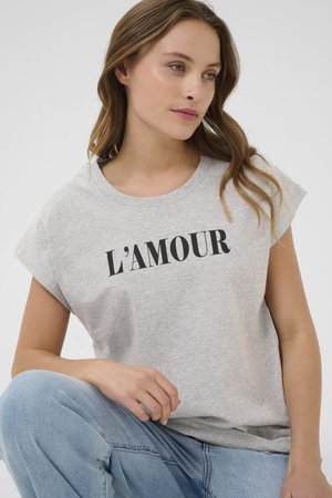 Kvinde med lyst brunt hår iført grå ærmeløs top med "L'AMOUR" i sorte bogstaver og lyseblå jeans, siddende med hånden hvilende på knæet.