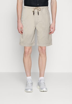 Mann in beigen Kordelzugshorts, weißen Socken und grauen Sneakers, der vor einem schlichten weißen Hintergrund steht.