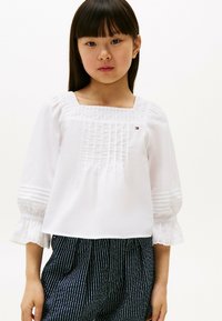 Blusa bianca con scollatura quadrata, pannelli testurizzati e maniche a sbuffo. Indossata con pantaloni a righe blu e bianchi. Presenta dettagli con logo.