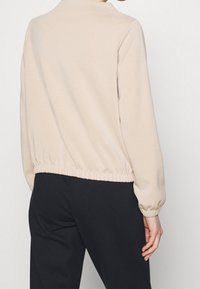 Beżowa bluza z elastycznym dołem i mankietami, o prostym designie. Noszona na ciemnych spodniach, ukazująca czyste linie i miękką teksturę.
