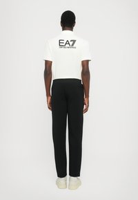 Polo shirt blanc avec logo EA7 noir dans le dos, associé à un pantalon de jogging noir et des baskets claires. Design simple et sportif.