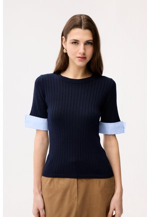 Kobieta o długich brązowych włosach, ubrana w dopasowany granatowy prążkowany sweter z jasnoniebieskimi podwiniętymi rękawami oraz beżowe spodnie, stojąca na białym tle.