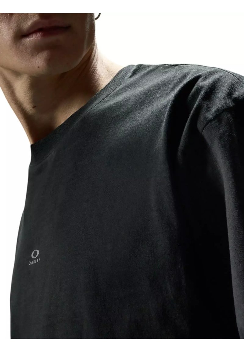 Homme portant un t-shirt noir uni Oakley, vue recadrée montrant le cou, l'épaule et une partie du visage sur fond blanc.