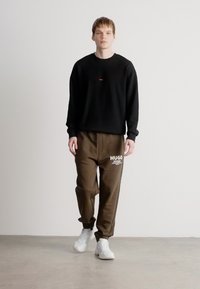 Μαύρη φούτερ μπλούζα με μικρό κόκκινο λογότυπο, σε συνδυασμό με χαλαρές ελαιοπράσινες φόρμες που έχουν το "HUGO" σε λευκό χρώμα. Οι λευκές αθλητικές παπούτσια ολοκληρώνουν το σύνολο.