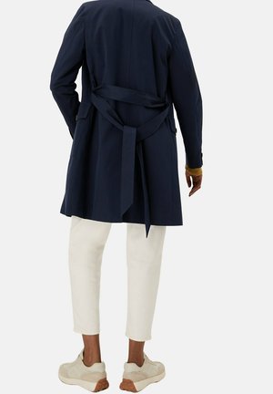 Donkerblauwe trenchcoat met een strikceintuur, lange mouwen en een knielengte snit. Gecombineerd met witte cropped broek en lichtgekleurde sneakers.