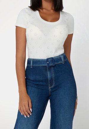 Kvinde iført en tætsiddende hvid top med korte ærmer med sølvfarvede "G"-mønstre og højttaljede blå jeans med et matchende bælte i denim.