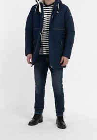 Navy padded jacket with a hood, white fleece lining, front zipper, pockets, worn over a black-and-white striped shirt and blue jeans.