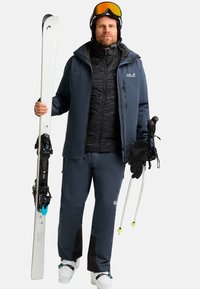 Attrezzatura da sci che include una giacca blu scuro, un gilet nero imbottito, pantaloni scuri, sci bianchi e bastoncini con dettagli verdi. Include guanti neri e occhiali da sci.