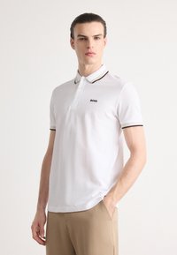 Hvit polo skjorte laget av bomull, med en to-knaps placket, svart og gullstripet krage og mansjetter, samt en liten svart logo på brystet.