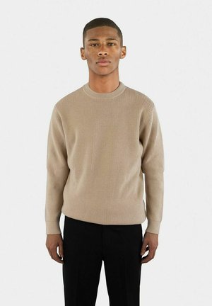 Beige ribbad stickad tröja med rund halsringning och långa ärmar, i kombination med svarta byxor, bärs av en modell framför en vit bakgrund.