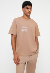 Bežinis medvilninis marškinėliai su trumpomis rankovėmis, apvaliu iškirpimu, priekyje su baltu "Calvin Klein" logotipu. Lygus paviršius, laisvas siluetas.