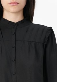 Blusa nera con bottoni frontali, colletto arricciato e dettagli plissettati sulle spalle. Il tessuto ha una consistenza morbida e liscia.