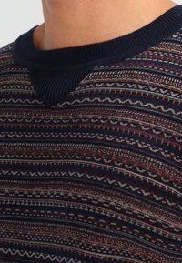 Marinblå tröja med strukturerad stickning och geometriska mönster i röd och vit, ribbad halsringning och en subtil v-formad accent vid kragen.