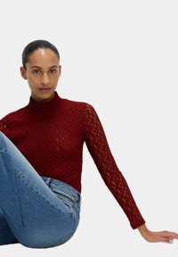 Top in pizzo borgogna con colletto alto e maniche lunghe. Le maniche presentano un design semi-trasparente, abbinato a jeans in denim blu.