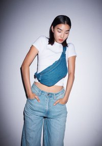 Borsa a tracolla in denim di un blu brillante, caratterizzata da una forma curvata, chiusura con zip e tracolla regolabile. Indossata sopra una maglietta bianca e jeans chiari.