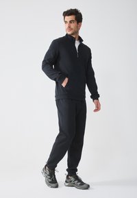 Hanorac negru cu fermoar, cu guler înalt și buzunar tip kangaroo, asortat cu pantaloni corespunzători. Modelul poartă încălțăminte sport gri și neagră.