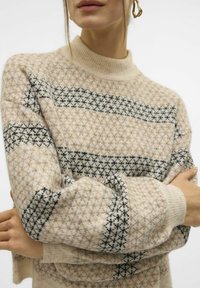 Beige trui met een hoge hals. Beschikt over een geometrisch patroon in donkerblauwe en zwarte strepen. Zachte gebreide textuur. Losse pasvorm.