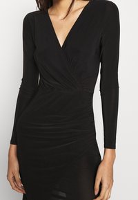 Černé večerní šaty s dlouhým rukávem, vyrobené z hladké látky, s hlubokým výstřihem do V, nařasenými detaily a těsným střihem.