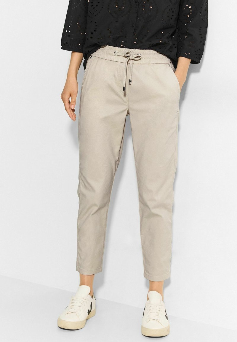 Ljusbeige tapered byxor med elastisk dragsko i midjan, sidofickor och en mjuk bomullstextur, kombinerat med vita sneakers.