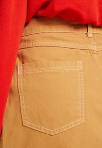 Pantalon en coton beige avec une poche arrière, ornés de coutures blanches contrastantes. Un pull rouge est partiellement visible au-dessus.
