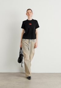 Svart bomull t-skjorte med rødt logo, parret med beige vide bukser. Modellen bærer en svart håndveske, sko med spiss tå, nøytral bakgrunn.