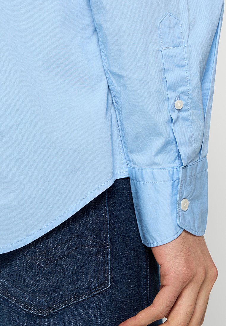 Camicia azzurra a maniche lunghe con colletto a punta, polsini con bottoni e orlo leggermente curvato, abbinata a pantaloni in denim scuro.