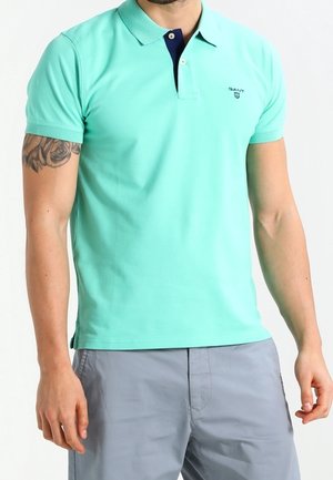 Man draagt een mintgroen poloshirt met korte mouwen, een donkerblauwe knopenbies en lichtgrijze korte broek, met een zichtbare tatoeage op zijn arm.