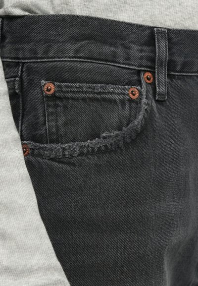 Schwarze Denim-Jeans mit einer verblassten Textur, ausgefransten Taschenkanten und braunen Nieten. Begleitet von einem hellgrauen texturierten Stoff.