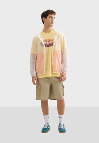 Coupe-vent transparent avec capuche, orné d'accents pastel orange et rose, porté sur un t-shirt graphique jaune, un short cargo et des baskets bleues.