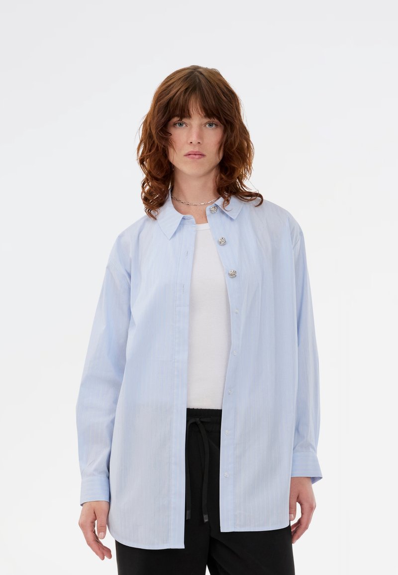 Hele sinine nööpidega särk, millel on triibuline muster, pikad varrukad ja lõdvestatud lõige. Omab hõbedaseid nööpe ja klassikalist kaelust.